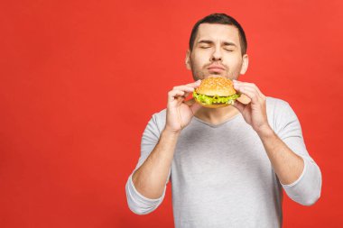 Hamburger tutan genç bir adam. Öğrenci fast food yiyor. Burger yararlı bir yemek değildir. Çok aç bir adam. Diyet konsepti. Kırmızı arkaplan üzerinde izole edilmiş.