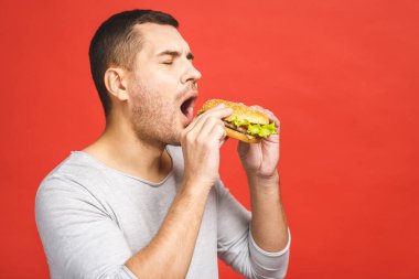 Hamburger tutan genç bir adam. Öğrenci fast food yiyor. Burger yararlı bir yemek değildir. Çok aç bir adam. Diyet konsepti. Kırmızı arkaplan üzerinde izole edilmiş.