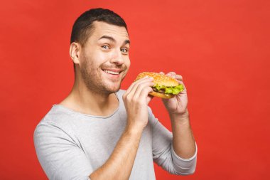 Hamburger tutan genç bir adam. Öğrenci fast food yiyor. Burger yararlı bir yemek değildir. Çok aç bir adam. Diyet konsepti. Kırmızı arkaplan üzerinde izole edilmiş.