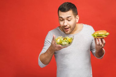 Salata ve hamburger, sağlıklı ve abur cubur arasında seçim yapmayı düşünen bir adam. Diyet konsepti. Sağlıklı ve sağlıksız yemek arasında seçim yapmak. Organik gıdalar fast food 'a karşı, kırmızı üzerine izole edilmiş.. 