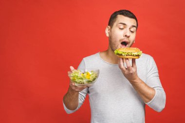 Salata ve hamburger, sağlıklı ve abur cubur arasında seçim yapmayı düşünen bir adam. Diyet konsepti. Sağlıklı ve sağlıksız yemek arasında seçim yapmak. Organik gıdalar fast food 'a karşı, kırmızı üzerine izole edilmiş.. 