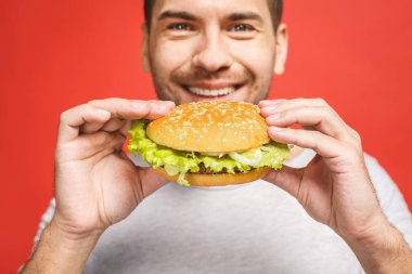 Hamburger tutan genç bir adam. Öğrenci fast food yiyor. Burger yararlı bir yemek değildir. Çok aç bir adam. Diyet konsepti. Kırmızı arkaplan üzerinde izole edilmiş.