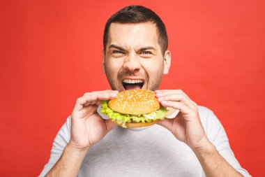 Hamburger tutan genç bir adam. Öğrenci fast food yiyor. Burger yararlı bir yemek değildir. Çok aç bir adam. Diyet konsepti. Kırmızı arkaplan üzerinde izole edilmiş.