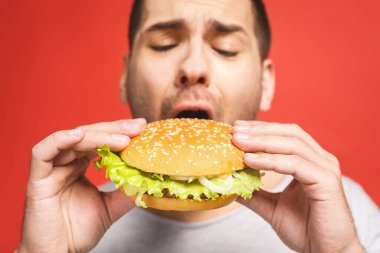 Hamburger tutan genç bir adam. Öğrenci fast food yiyor. Burger yararlı bir yemek değildir. Çok aç bir adam. Diyet konsepti. Kırmızı arkaplan üzerinde izole edilmiş.