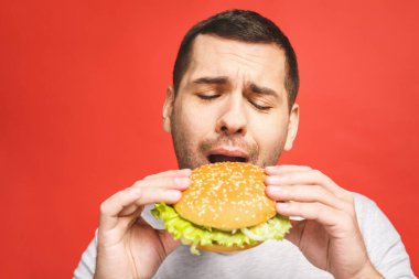 Hamburger tutan genç bir adam. Öğrenci fast food yiyor. Burger yararlı bir yemek değildir. Çok aç bir adam. Diyet konsepti. Kırmızı arkaplan üzerinde izole edilmiş.