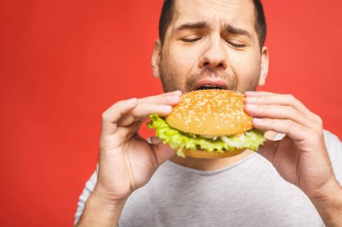 Hamburger tutan genç bir adam. Öğrenci fast food yiyor. Burger yararlı bir yemek değildir. Çok aç bir adam. Diyet konsepti. Kırmızı arkaplan üzerinde izole edilmiş.