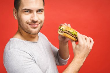 Hamburger tutan genç bir adam. Öğrenci fast food yiyor. Burger yararlı bir yemek değildir. Çok aç bir adam. Diyet konsepti. Kırmızı arkaplan üzerinde izole edilmiş.