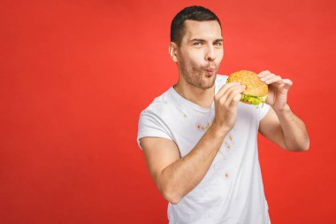 Abur cubur yiyen komik, aç sakallı adam. Heyecanlı genç adam aç gözlülükle kırmızı arka planda izole edilmiş hamburgerleri yiyor..