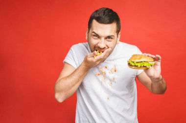 Abur cubur yiyen komik, aç sakallı adam. Heyecanlı genç adam aç gözlülükle kırmızı arka planda izole edilmiş hamburgerleri yiyor..