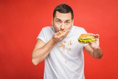 Abur cubur yiyen komik, aç sakallı adam. Heyecanlı genç adam aç gözlülükle kırmızı arka planda izole edilmiş hamburgerleri yiyor..