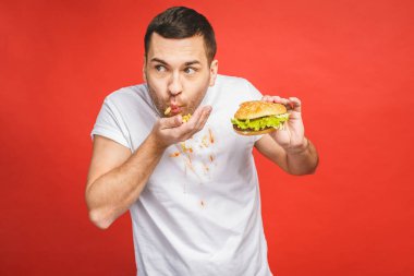 Abur cubur yiyen komik, aç sakallı adam. Heyecanlı genç adam aç gözlülükle kırmızı arka planda izole edilmiş hamburgerleri yiyor..