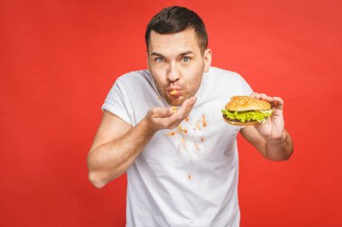 Abur cubur yiyen komik, aç sakallı adam. Heyecanlı genç adam aç gözlülükle kırmızı arka planda izole edilmiş hamburgerleri yiyor..