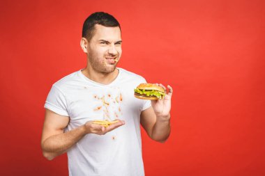 Abur cubur yiyen komik, aç sakallı adam. Heyecanlı genç adam aç gözlülükle kırmızı arka planda izole edilmiş hamburgerleri yiyor..