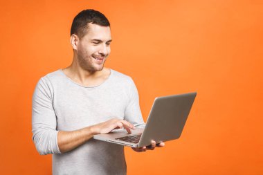 Kendine güvenen bir iş uzmanı. Dizüstü bilgisayarı olan ve turuncu arka planda dururken gülümseyen kendinden emin, genç ve yakışıklı bir adam..