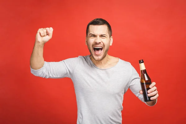 stock image A young guy with a beard isolated on a red background holds a bottle of beer. Watching football match.