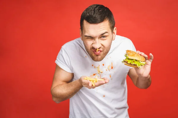Funny hungry bearded man eating junk food. Excited young man greedily eating hamburgers isolated on red background.