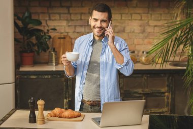 Sabah haberleri! Yakışıklı genç adam evde ev mutfağında laptopuyla kahve içiyor, gülümsüyor, telefon kullanıyor..