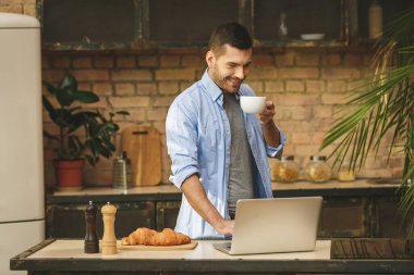 Sabah haberleri! Yakışıklı genç adam evde ev mutfağında laptopuyla kahve içiyor, gülümsüyor..