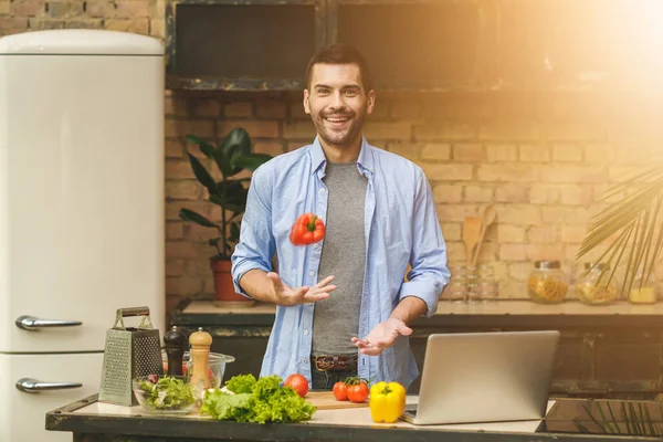 とってもおいしい ロフトキッチンで自宅でサラダを準備し 笑顔カジュアルな幸せな若い男 ノートパソコンを使う — ストック写真