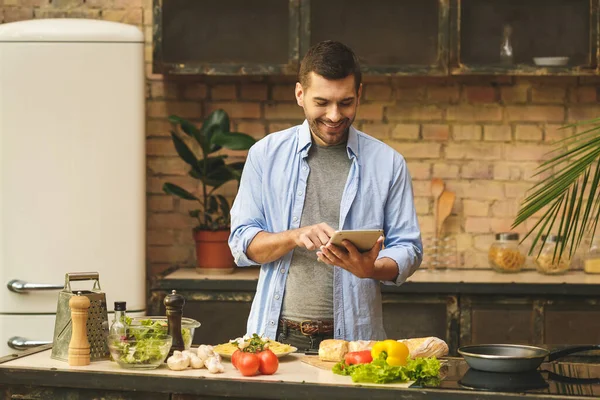 Man preparing delicious and healthy food in the home kitchen on a sunny day. Using tablet computer.