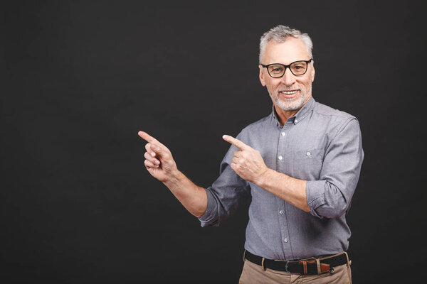 Aged senior man wearing glasses isolated against black background amazed and smiling to the camera while presenting with hand and pointing with finger.