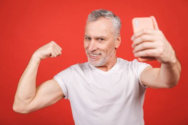 Kaslarını gösteren, akıllı telefondan selfie çeken, kırmızı arka planda izole edilmiş mutlu yaşlı sakallı bir adamın portresi. Spor ve sağlıklı konsept. 