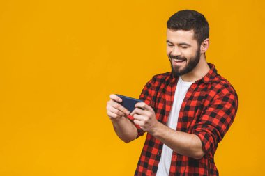 Ekose gömlekli heyecanlı sakallı adam sarı arka planda izole bir şekilde akıllı telefonuyla oynuyor..