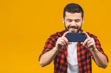 Ekose gömlekli heyecanlı sakallı adam sarı arka planda izole bir şekilde akıllı telefonuyla oynuyor..