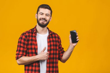 Gülümseyen çekici bir adamın portresi boş ekranlı cep telefonu tutuyor ve sarı arkaplanda izole edilmiş baş parmak hareketlerini gösteriyor..