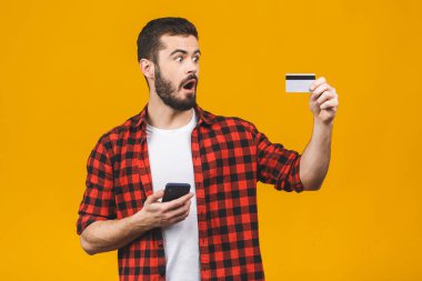 Akıllı telefon kullanan genç bir adam sarı arka planda izole edilmiş. Şaşırmış bir yüzle korkmuş, korku ifadesi ve kredi kartıyla heyecanlanmış, alışveriş yapıyor.. 