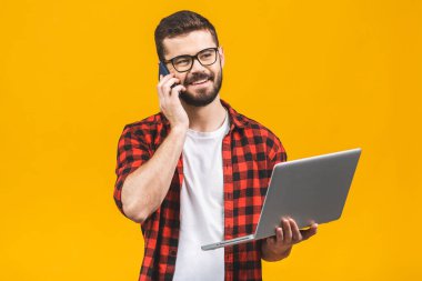 Sırıtkan genç iş adamı dizüstü bilgisayarla oturmuş akıllı telefonuyla arıyor..