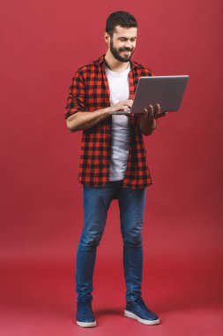 İşine odaklan. Neşeli genç bir adamın tam boy portresi. Kırmızı arka planda dikilirken dizüstü bilgisayarda çalışan kendine güvenen yakışıklı adam..