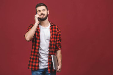 Kırmızı arka planda izole edilmiş cep telefonuyla konuşurken sıradan bir dizüstü bilgisayardaki mutlu gülümseyen iş adamının portresi..
