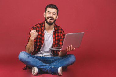 Laptop kullanan mutlu bir gencin portresi ve kırmızı arkaplanda izole edilmiş başarıyı kutlamak..