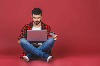 Kırmızı arka planda bacak bacak üstüne atmış, dizüstü bilgisayar kullanan, kredi kartını gösteren neşeli genç bir adamın tam uzunluğu..