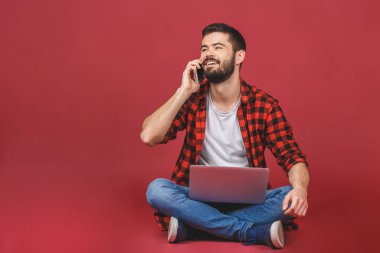 Yerde dizüstü bilgisayarı olan ve telefonda konuşan bir adam var. Kırmızı arkaplanda izole edilmiş.
