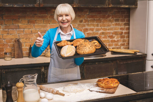 Portrait of attractive smiling happy senior aged woman is cooking on kitchen. Grandmother making tasty baking. Thumbs up.