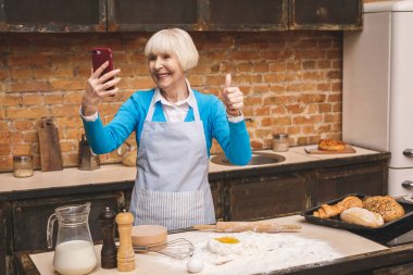 Selfie konsepti. Yaşlı çekici bir kadının portresi mutfakta yemek pişiriyor. Büyükannem lezzetli pişirme yapıyor. Başparmaklar yukarı..