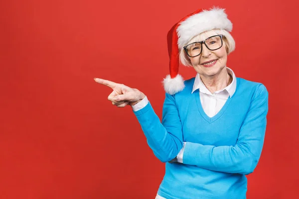 Old senior hispanic woman wearing christmas hat over isolated red background smiling cheerful presenting and pointing with palm of hand looking at the camera.