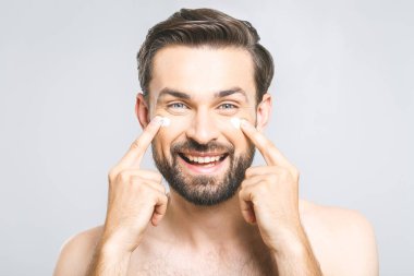 Cilt bakımı. Yakışıklı, mutlu, üstsüz bir adam yüzüne krem sürüyor ve gri arka planda durup kameraya bakarken gülümseyerek kendine bakıyor. Yakın plan. Metin için boşluk.
