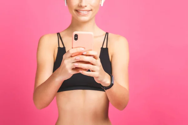 Close-up portrait of joyful beautiful fitness woman listening music by smartphone and dancing isolated over pink background. Using phone.