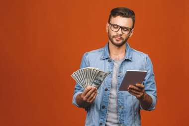 Turuncu duvar arkasında yalnız poz veren heyecanlı, genç sakallı adamın görüntüsü. Elinde para ve tablet bilgisayarla birinci sınıf bir jest yapıyor..