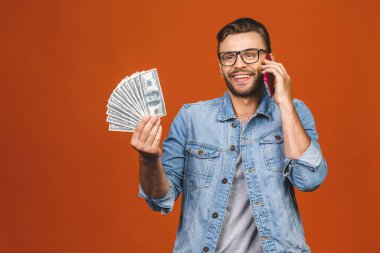Havalı tişörtlü, elinde bir sürü parayla turuncu duvardan izole edilmiş bir telefon kullanan heyecanlı bir adam.