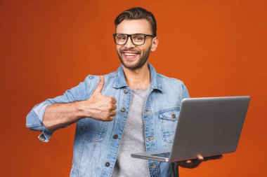 Portakal rengi arka planda, reklam için fotokopi alanı olan, dizüstü bilgisayarı olan ve mutlu bir gülümsemeyle kameraya bakan sıradan bir genç adam portresi. Başparmaklar yukarı..