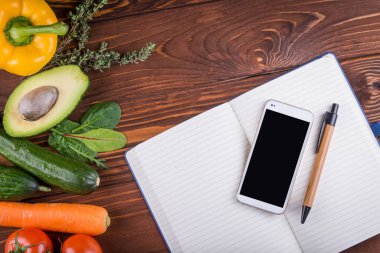 Taze organik sebzeler ve meyveler. Biber, domates, avokado, boş bir defter, ahşap arka planda telefon ve kalem. Sağlıklı yiyecekler ve sağlıklı yaşam konsepti. Üst görünüm