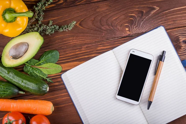 Taze organik sebzeler ve meyveler. Biber, domates, avokado, boş bir defter, ahşap arka planda telefon ve kalem. Sağlıklı yiyecekler ve sağlıklı yaşam konsepti. Üst görünüm