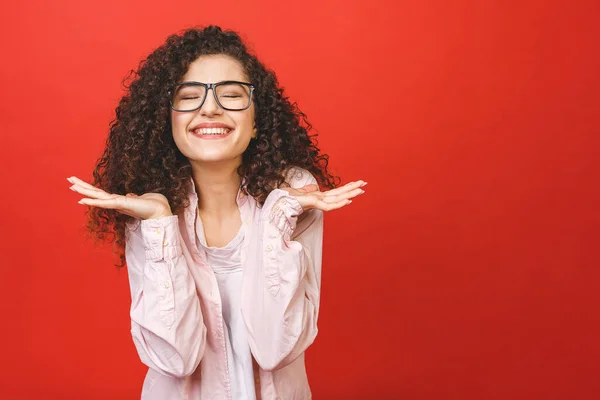 Happy cheerful young woman with curly hair rejoicing at positive news or birthday gift, looking at camera with joyful and charming smile. Student girl relaxing after college isolated over red background.