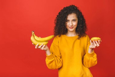 Kafası karışık, üzgün kıvırcık kadın sağlıklı ve sağlıksız yemek arasında seçim yapamaz. Elinde meyveli çörek, kırmızı arka planda soyutlanmış..
