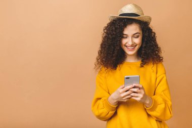 Bej arka planda izole edilmiş akıllı telefonu tutan, gülümseyen, kıvırcık öğrenci bir kadının portresi..