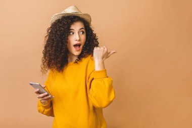 Neşeli, kıvırcık bir kız öğrencisinin portresi cep telefonunu tutuyor ve bej arka planda izole edilmiş bir şekilde işaret ediyor..